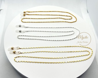 May include: Three eyeglass chains in gold and silver tones are displayed on a white surface. The chains have different designs, including a beaded chain, a rectangular link chain, and a chain with small square links. The chains are designed to hold eyeglasses.