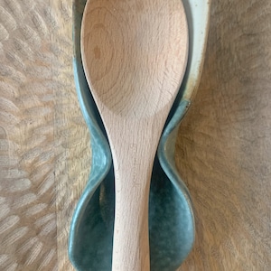 May include: A ceramic spoon rest with a green and white glaze. The rest is shaped like a teardrop and has a curved indentation to hold a spoon. A wooden spoon is resting in the indentation.