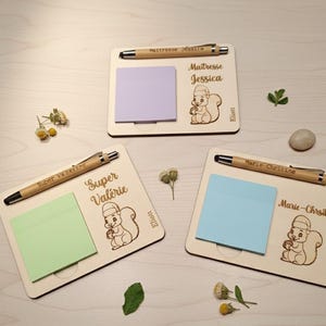 Op de afbeelding: Drie rechthoekige houten memoborden, elk voorzien van een bamboe pen, een vierkant memoblaadje in lichtpaars, lichtgroen of lichtblauw, en een lasergegraveerde eekhoornillustratie. Elk bord is gepersonaliseerd met namen zoals Maitresse Jessica, Super Valérie, Marie-Christine en de naam Eliott.