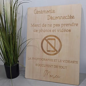 Peut inclure: Panneau en bois clair avec texte gravé : "Cérémonie Déconnectée", un symbole de téléphone barré et "Merci de ne pas prendre de photos et vidéos". Une plante en pot noir est visible à gauche.
