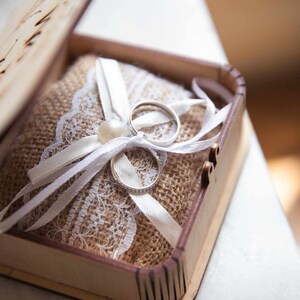 Peut inclure: Un écrin en bois contenant deux alliances en argent sur un coussin de toile de jute et de dentelle. Des rubans de satin blancs et une perle ornent les anneaux, pour une présentation élégante.