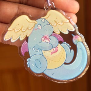 Puede incluir: Un llavero acrílico transparente con un dragón de dibujos animados abrazando a una criatura más pequeña. El dragón es azul claro con alas amarillas y detalles morados. El llavero tiene un cierre morado.