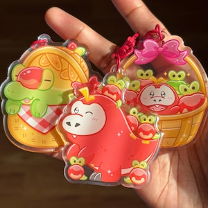 Puede incluir: Tres llaveros de acrílico transparente con personajes de dibujos animados. Uno muestra un caimán verde en un sándwich, otro una criatura roja con forma de dinosaurio y el último un cangrejo en una cesta. Cada llavero tiene un cordón rojo.