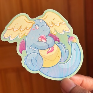 Könnte beinhalten: Ein Aufkleber mit einem Cartoon-Drachen in Blau-, Rosa- und Gelbtönen. Der Drache hat große Flügel und hält ein kleineres Wesen. Der Aufkleber hat einen hellgrünen Rand und einen regenbogenfarbenen Schwanz.
