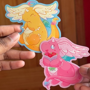 May include: Two colorful dragon stickers. One is yellow with orange accents, a halo, and wings. The other is pink, holding a basket of strawberries, with a halo and wings. Both have a cartoon style and are on a light blue background.