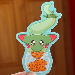 Puede incluir: Una pegatina caprichosa que presenta una criatura de dibujos animados con un cuerpo verde y detalles rojos, sosteniendo un tarro lleno de galletas con chispas de chocolate. La pegatina tiene un borde azul claro y un diseño lindo y juguetón.