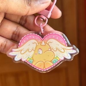Puede incluir: Un llavero rosa con un dije en forma de corazón con un unicornio de dibujos animados con alas. El dije tiene un cuerpo amarillo, detalles en rosa y un fondo de arcoíris. El llavero tiene un cierre rosa.