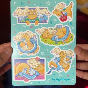 May include: A sheet of stickers featuring cartoon dragons in various poses. The dragons are primarily yellow and blue, with some green and pink accents. The stickers have a light blue background with decorative stars and hearts. Text on the stickers reads "By Appletunpiee".
