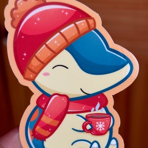 Puede incluir: Una pegatina de dibujos animados de un ping&uuml;ino con un gorro de invierno rojo y naranja y una bufanda, sosteniendo una taza roja con un dise&ntilde;o de copo de nieve. El ping&uuml;ino sonr&iacute;e y tiene una taza humeante de chocolate caliente.