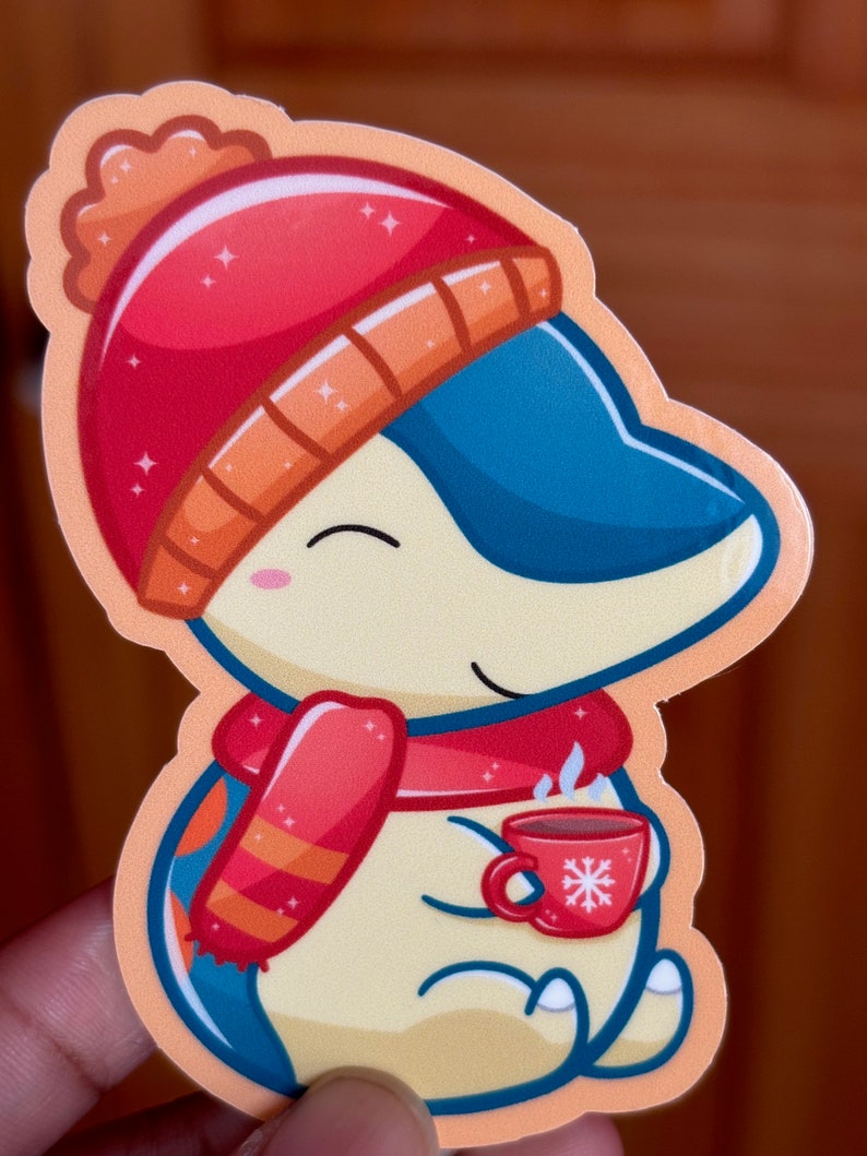 Puede incluir: Una pegatina de dibujos animados de un ping&uuml;ino con un gorro de invierno rojo y naranja y una bufanda, sosteniendo una taza roja con un dise&ntilde;o de copo de nieve. El ping&uuml;ino sonr&iacute;e y tiene una taza humeante de chocolate caliente.