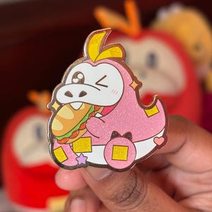 Puede incluir: Un pin de esmalte rosa con un cocodrilo de dibujos animados comiendo un sándwich. El pin tiene detalles de purpurina, detalles dorados y una expresión guiñando un ojo. El pin está en la mano de una persona.