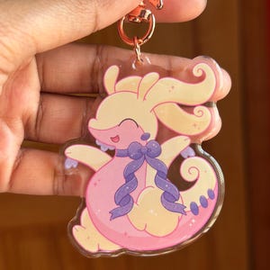 Peut inclure: Un porte-clés en acrylique transparent représentant un personnage de dessin animé aux tons rose et jaune, avec un nœud et des rubans violets. Le porte-clés a un fermoir en forme d'étoile en or rose.