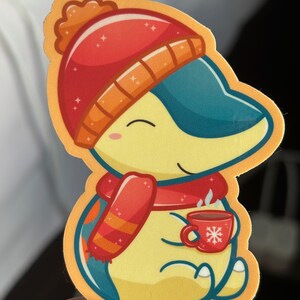 Puede incluir: Un personaje de dibujos animados amarillo que lleva un gorro y una bufanda rojos y naranjas, sosteniendo una taza roja con un dise&ntilde;o de copo de nieve. El personaje tiene una cola azul y sonr&iacute;e.