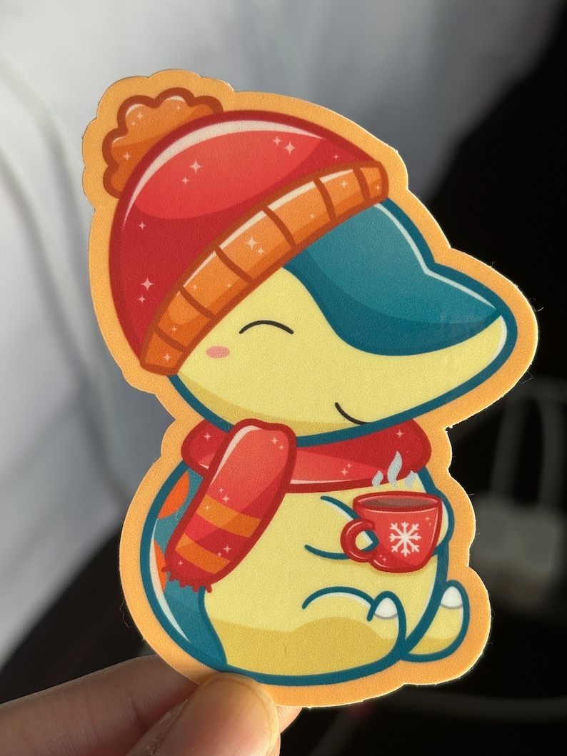 Puede incluir: Un personaje de dibujos animados amarillo que lleva un gorro y una bufanda rojos y naranjas, sosteniendo una taza roja con un dise&ntilde;o de copo de nieve. El personaje tiene una cola azul y sonr&iacute;e.