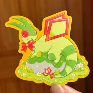 Puede incluir: Una pegatina troquelada con un dragón de dibujos animados en tonos verdes, con detalles rojos en los ojos, el lazo y las formas de alas. El dragón descansa sobre una roca gris, rodeado de hierba verde y pequeñas flores, en un borde amarillo.