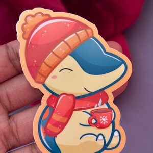 Puede incluir: Un ping&uuml;ino amarillo de dibujos animados que lleva un gorro rojo y naranja, una bufanda roja y sostiene una taza roja con un dise&ntilde;o de copo de nieve. El ping&uuml;ino est&aacute; sentado y tiene una expresi&oacute;n feliz en su rostro.