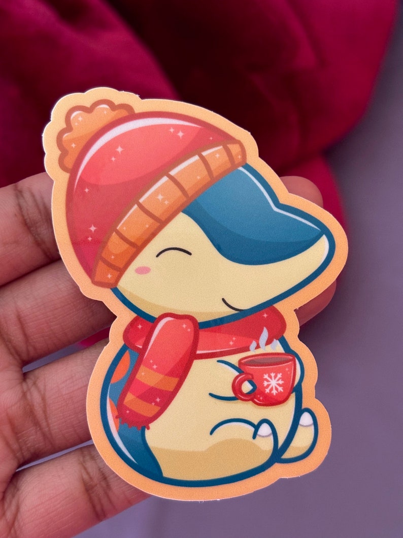 Puede incluir: Un ping&uuml;ino amarillo de dibujos animados que lleva un gorro rojo y naranja, una bufanda roja y sostiene una taza roja con un dise&ntilde;o de copo de nieve. El ping&uuml;ino est&aacute; sentado y tiene una expresi&oacute;n feliz en su rostro.