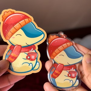 Puede incluir: Dos pegatinas de tem&aacute;tica invernal con un personaje de dibujos animados que lleva un gorro rojo y una bufanda, sosteniendo una taza de chocolate caliente. La pegatina m&aacute;s grande est&aacute; impresa en papel, mientras que la m&aacute;s peque&ntilde;a est&aacute; impresa en acr&iacute;lico transparente.