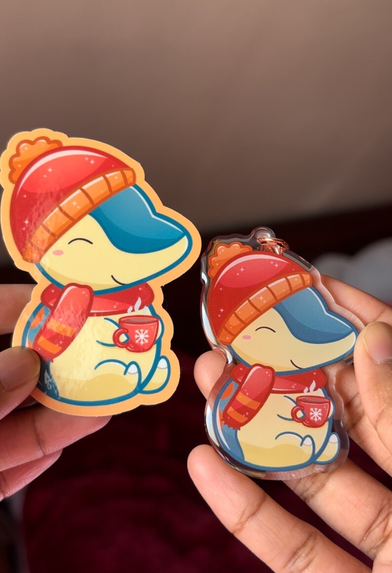 Puede incluir: Dos pegatinas de tem&aacute;tica invernal con un personaje de dibujos animados que lleva un gorro rojo y una bufanda, sosteniendo una taza de chocolate caliente. La pegatina m&aacute;s grande est&aacute; impresa en papel, mientras que la m&aacute;s peque&ntilde;a est&aacute; impresa en acr&iacute;lico transparente.