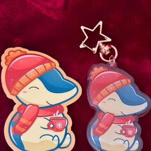 Puede incluir: Un llavero con tem&aacute;tica invernal que presenta un ping&uuml;ino de dibujos animados con un gorro rojo y blanco, una bufanda roja y sosteniendo una taza roja con un dise&ntilde;o de copo de nieve. El llavero est&aacute; hecho de acr&iacute;lico transparente y tiene un dije de estrella dorada.