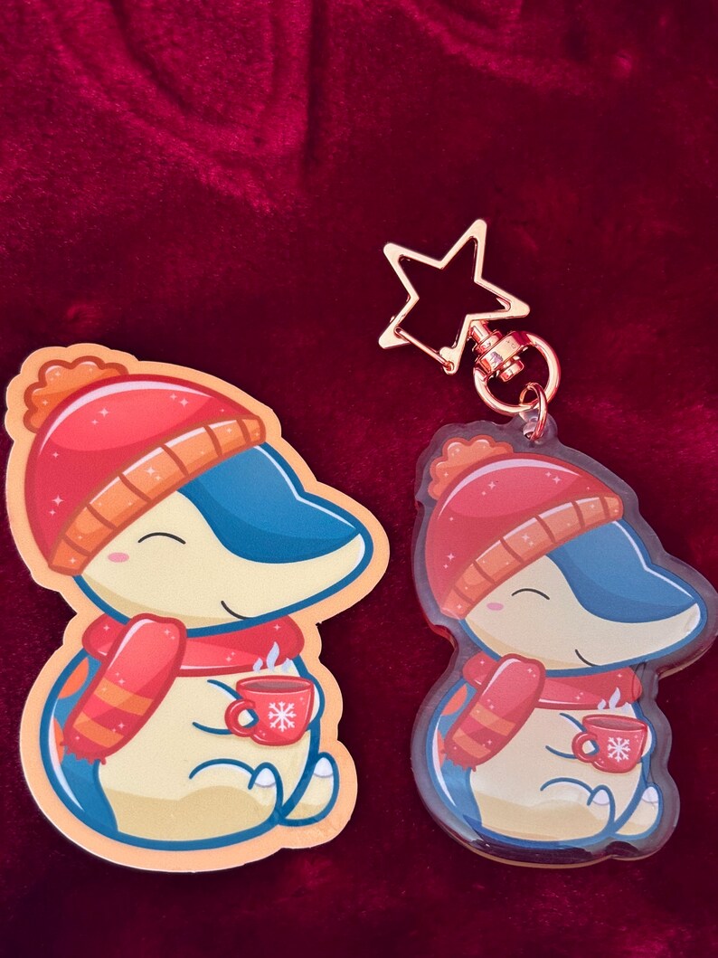 Puede incluir: Un llavero con tem&aacute;tica invernal que presenta un ping&uuml;ino de dibujos animados con un gorro rojo y blanco, una bufanda roja y sosteniendo una taza roja con un dise&ntilde;o de copo de nieve. El llavero est&aacute; hecho de acr&iacute;lico transparente y tiene un dije de estrella dorada.