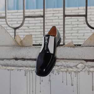 Puede incluir: Un zapato de cuero negro y blanco con una cremallera en el lateral. El zapato est&aacute; colgado de una pared de ladrillo con una rejilla met&aacute;lica azul detr&aacute;s. El texto "BIU CILI" y "ABOUT ASYMMETRIC CHIC" est&aacute; impreso debajo del zapato.