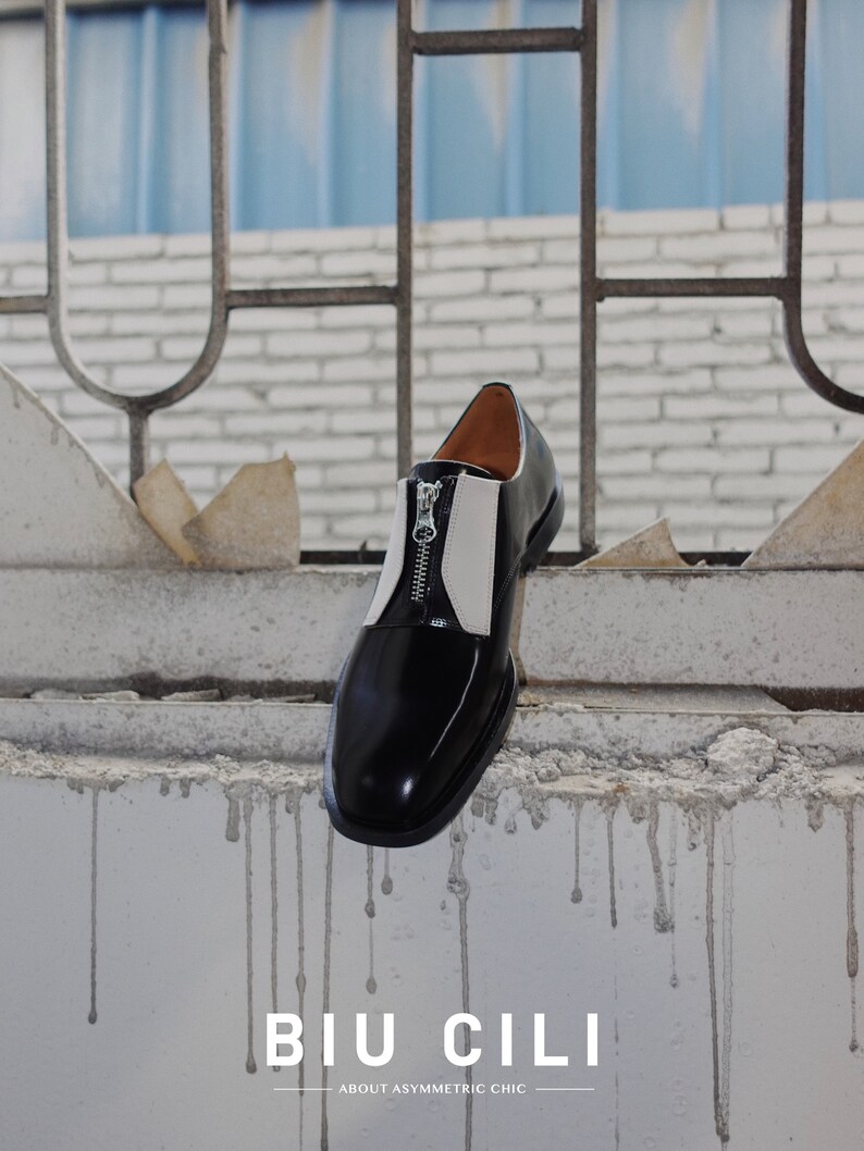 Puede incluir: Un zapato de cuero negro y blanco con una cremallera en el lateral. El zapato est&aacute; colgado de una pared de ladrillo con una rejilla met&aacute;lica azul detr&aacute;s. El texto "BIU CILI" y "ABOUT ASYMMETRIC CHIC" est&aacute; impreso debajo del zapato.