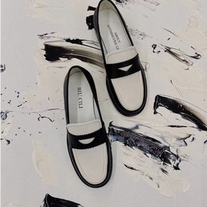 May include: A pair of black and white leather loafers with a contrasting white trim and a black leather strap across the toe. The loafers have the text "ABOUT  OF ASYMMETRIC" on one shoe and "BUCCLLI" on the other shoe.