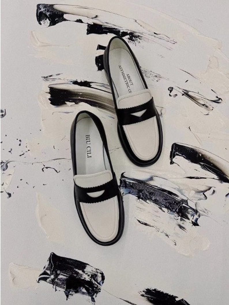 May include: A pair of black and white leather loafers with a contrasting white trim and a black leather strap across the toe. The loafers have the text "ABOUT  OF ASYMMETRIC" on one shoe and "BUCCLLI" on the other shoe.