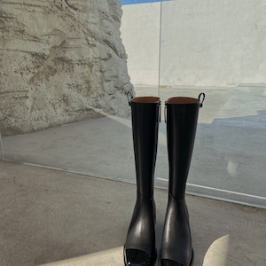 May include: A pair of black leather knee-high boots with a square toe and a shiny black cap toe.