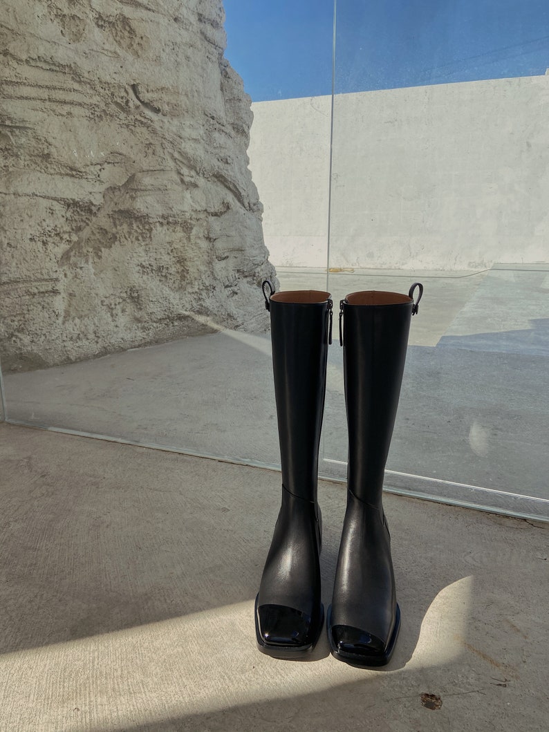 May include: A pair of black leather knee-high boots with a square toe and a shiny black cap toe.