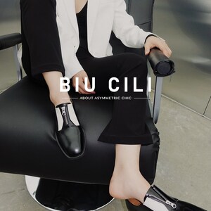 Puede incluir: Una persona con un blazer blanco y pantalones negros se sienta en una silla. Lleva zapatos de cuero negro con un detalle de cremallera blanca. Los zapatos tienen una punta puntiaguda y un tac&oacute;n grueso. El texto "BIU CILI" y "ABOUT ASYMMETRIC CHIC" es visible en la silla.