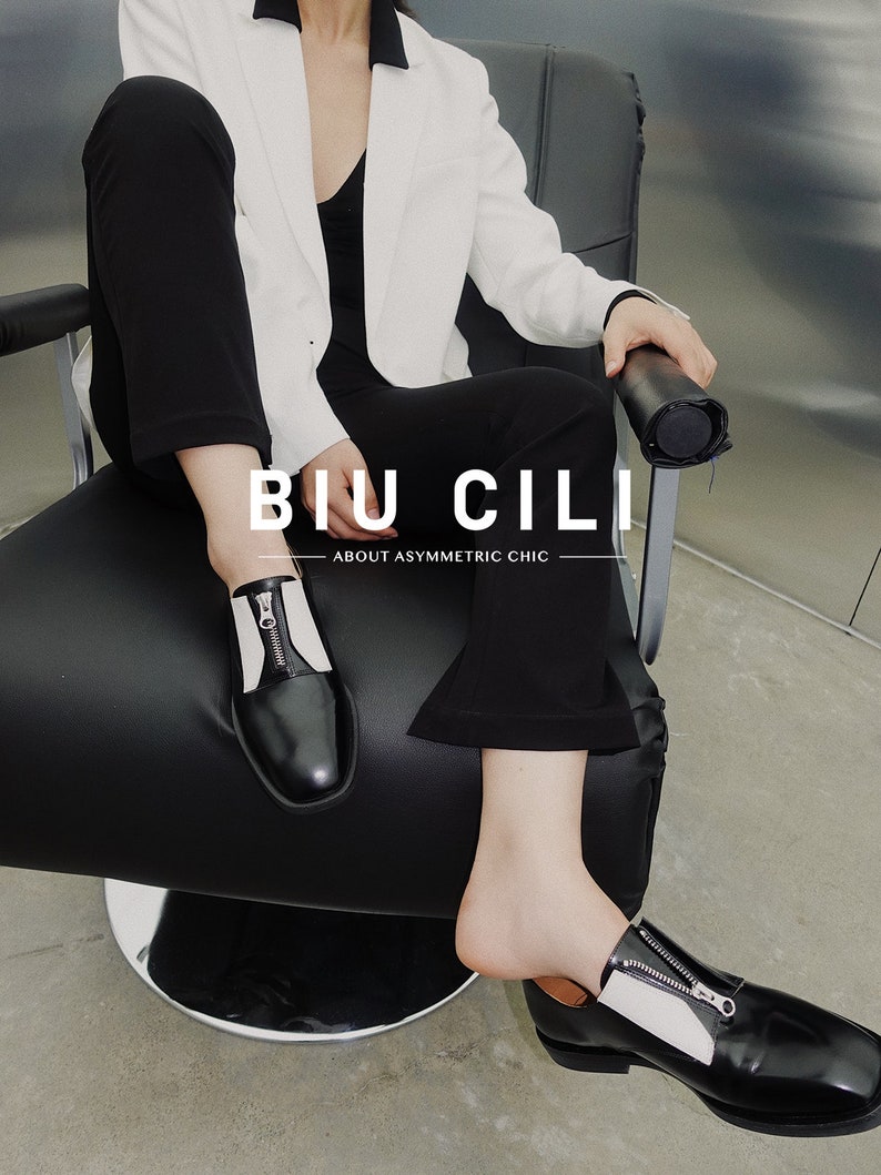 Puede incluir: Una persona con un blazer blanco y pantalones negros se sienta en una silla. Lleva zapatos de cuero negro con un detalle de cremallera blanca. Los zapatos tienen una punta puntiaguda y un tac&oacute;n grueso. El texto "BIU CILI" y "ABOUT ASYMMETRIC CHIC" es visible en la silla.