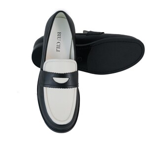 May include: Black and white leather loafers with a contrasting color design. The loafers have a black leather upper with a white leather toe and a black leather heel. The loafers have a black leather strap across the top of the foot with a silver buckle. The loafers have a black leather sole. The loafers have the text "BIU CILI" printed on the inside of the shoe.