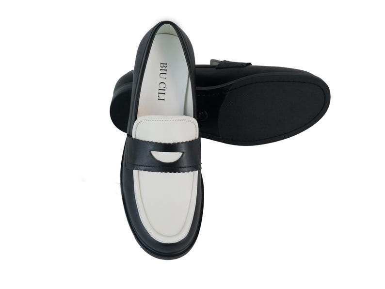 May include: Black and white leather loafers with a contrasting color design. The loafers have a black leather upper with a white leather toe and a black leather heel. The loafers have a black leather strap across the top of the foot with a silver buckle. The loafers have a black leather sole. The loafers have the text "BIU CILI" printed on the inside of the shoe.