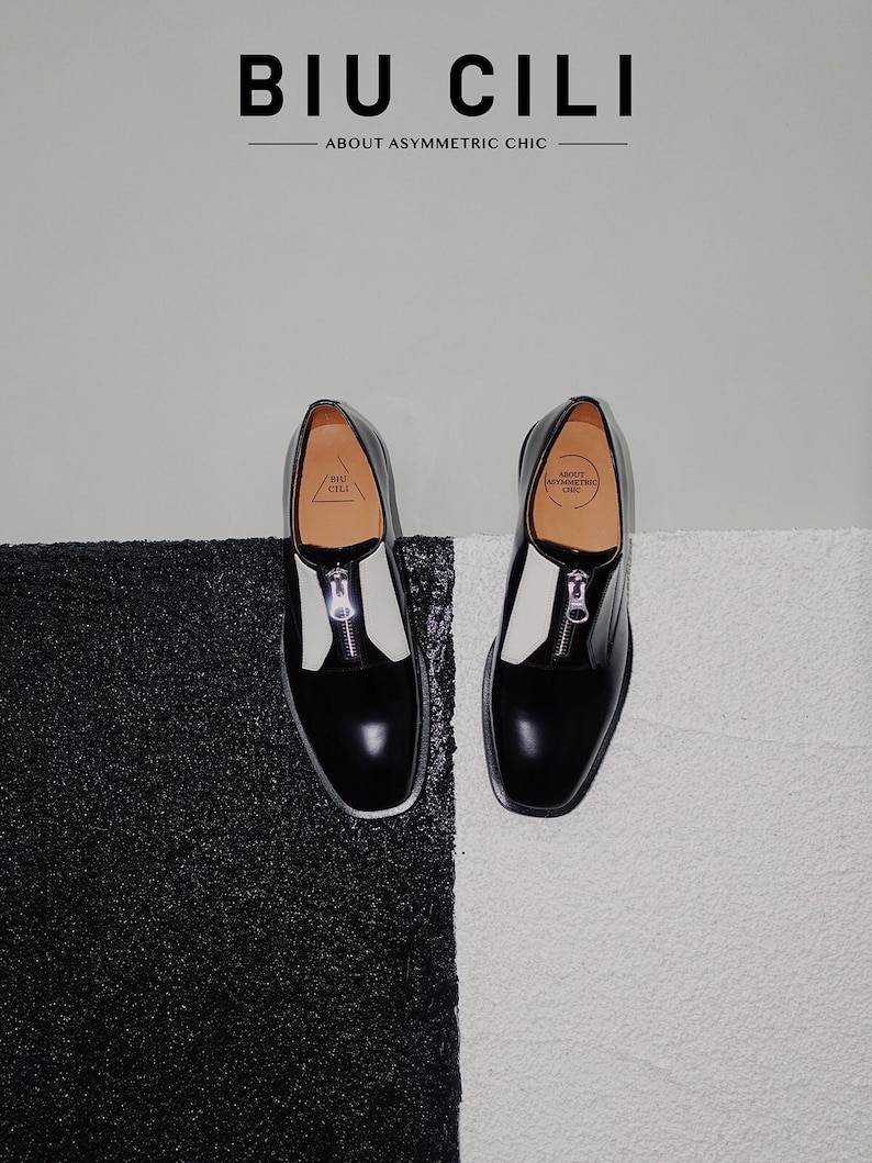 Puede incluir: Un par de zapatos de cuero negro y blanco con un detalle de cremallera. Los zapatos tienen un panel de cuero blanco en la parte superior y un panel de cuero negro en la parte inferior. Los zapatos est&aacute;n sobre un fondo blanco y negro. El texto "BIU CILI ABOUT ASYMMETRIC CHIC" es visible sobre los zapatos.
