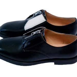Puede incluir: Un par de zapatos de cuero negro con un detalle de cremallera. Los zapatos tienen un panel de cuero blanco en la parte superior del zapato.