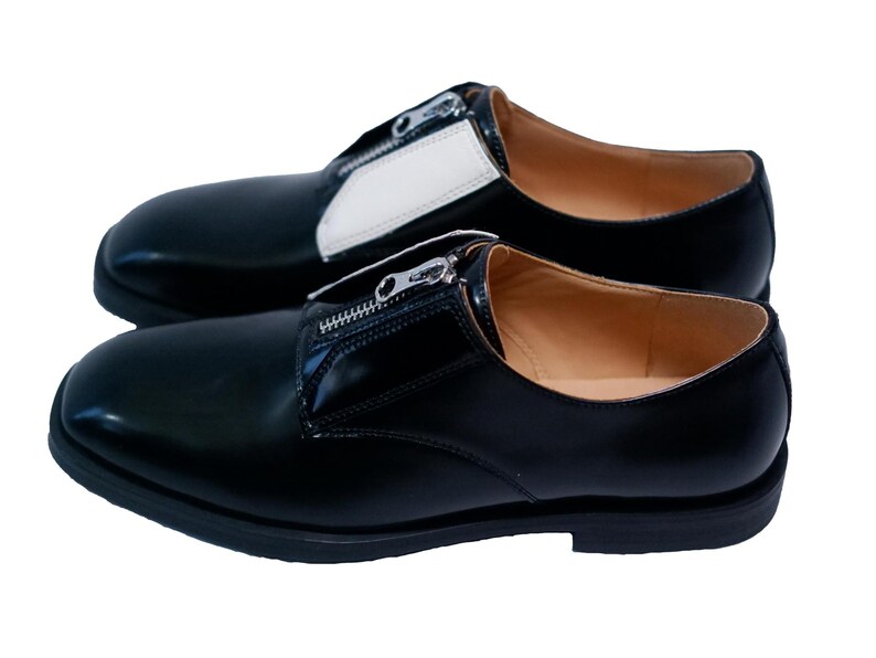 Puede incluir: Un par de zapatos de cuero negro con un detalle de cremallera. Los zapatos tienen un panel de cuero blanco en la parte superior del zapato.