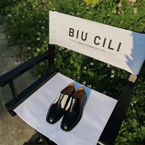 Puede incluir: Un par de zapatos de cuero negro con paneles blancos y detalles de cremallera en una silla de director blanca. La silla tiene un marco negro y un respaldo de lona blanca con el texto "BIU CILI - ABOUT ASYMMETRIC CHIC".