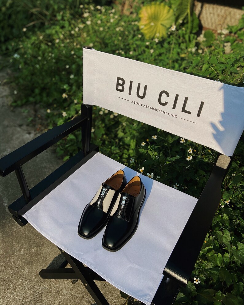 Puede incluir: Un par de zapatos de cuero negro con paneles blancos y detalles de cremallera en una silla de director blanca. La silla tiene un marco negro y un respaldo de lona blanca con el texto "BIU CILI - ABOUT ASYMMETRIC CHIC".