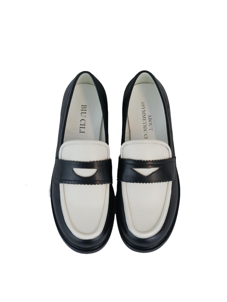 May include: A pair of black and white leather loafers with a contrasting color design. The loafers have a black leather upper with a white leather toe and heel. The text "BIU CILI" is printed on the inside of the left shoe and "ASYMMETRIC" is printed on the inside of the right shoe.