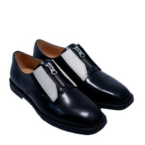 Puede incluir: Par de zapatos de cuero negro con paneles de cuero blanco y cremalleras plateadas. Los zapatos tienen una punta redonda y una suela gruesa.