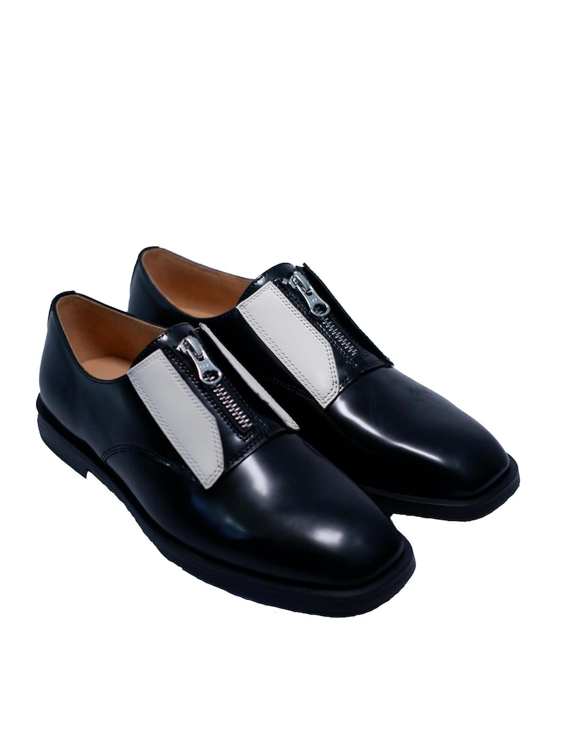 Puede incluir: Par de zapatos de cuero negro con paneles de cuero blanco y cremalleras plateadas. Los zapatos tienen una punta redonda y una suela gruesa.
