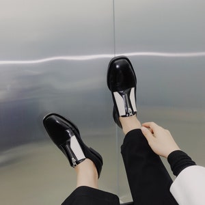 Puede incluir: Zapatos de cuero negro y blanco con detalle de cremallera. Los zapatos tienen una punta redonda y un tac&oacute;n bajo.