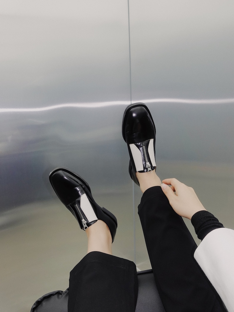 Puede incluir: Zapatos de cuero negro y blanco con detalle de cremallera. Los zapatos tienen una punta redonda y un tac&oacute;n bajo.