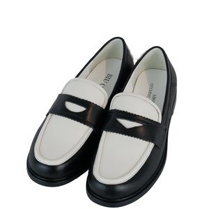 May include: A pair of black and white leather loafers with a contrasting color design. The loafers have a black leather upper with a white leather toe and heel. The loafers have a black leather strap across the vamp with a small buckle.