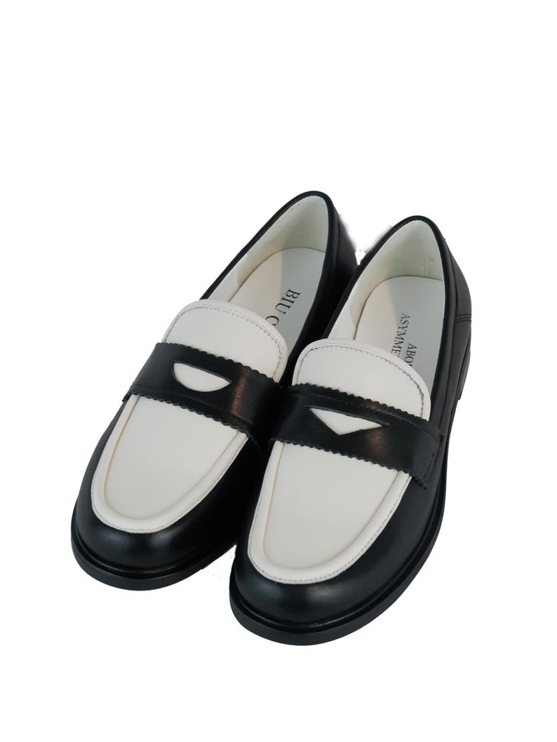May include: A pair of black and white leather loafers with a contrasting color design. The loafers have a black leather upper with a white leather toe and heel. The loafers have a black leather strap across the vamp with a small buckle.