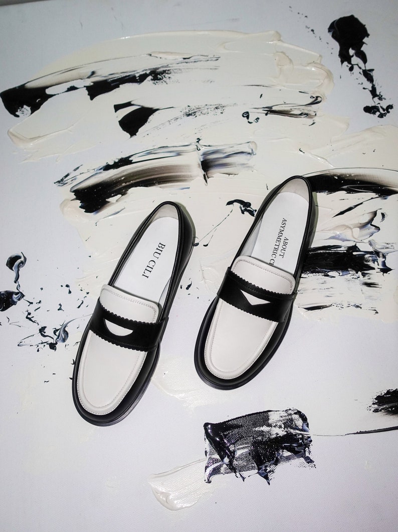 May include: A pair of black and white leather loafers with a black trim and a white sole. The shoes are on a white background with black paint splatters.