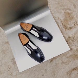 Puede incluir: Un par de zapatos de cuero negro con paneles blancos y un detalle de cremallera. Los zapatos est&aacute;n sobre una superficie plateada.