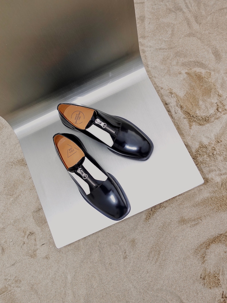 Puede incluir: Un par de zapatos de cuero negro con paneles blancos y un detalle de cremallera. Los zapatos est&aacute;n sobre una superficie plateada.